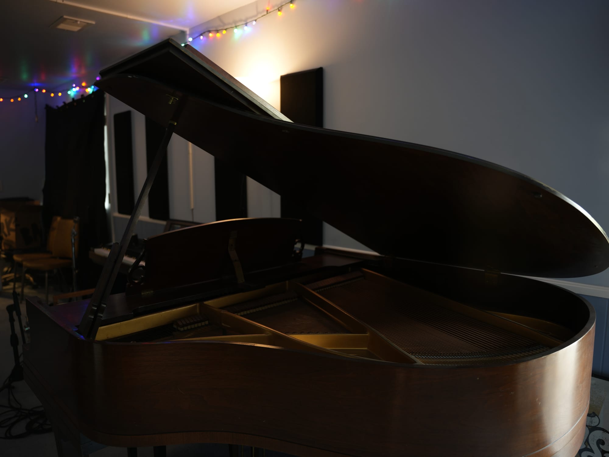A wooden baby grand piano with the lid open in the live room, lit by string lights.